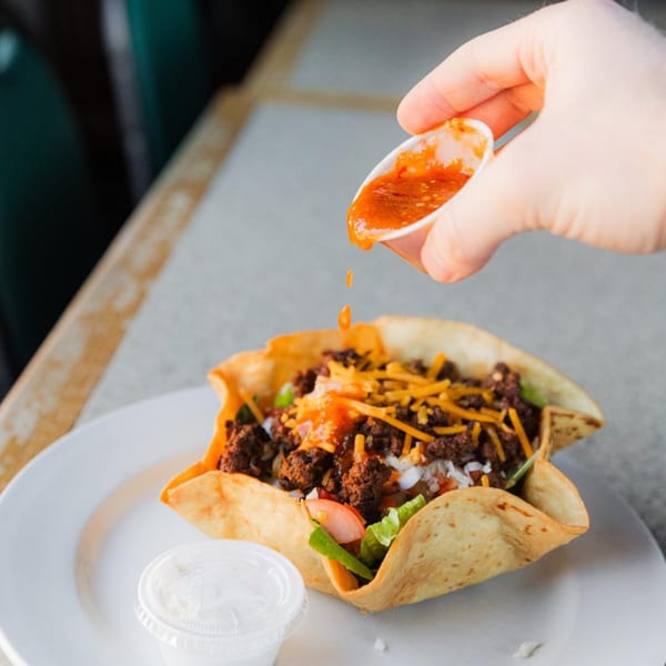 A hand pours salsa from a small cup onto a taco salad in a crispy shell.