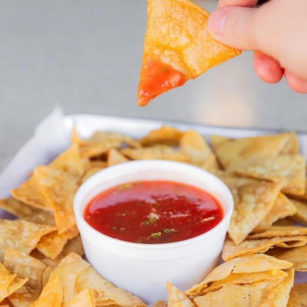 A hand dips a crispy tortilla chip into a white bowl of red salsa, surrounded by more chips.