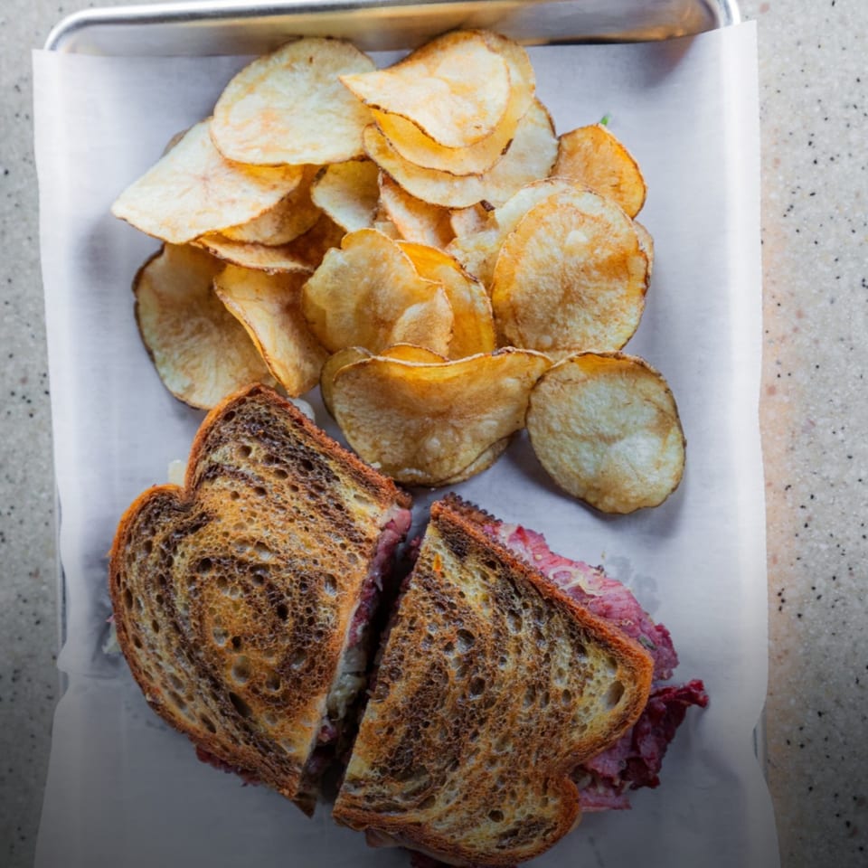 A Reuben sandwich cut in half, served with a pile of golden potato chips on a tray.