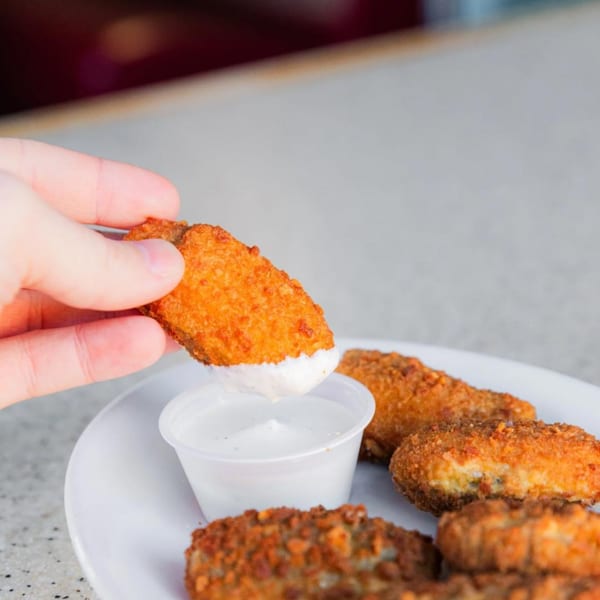 A hand dips a golden-brown fried pickle into a small cup of white dipping sauce on a plate.